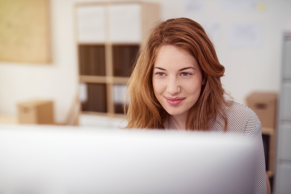 Attractive young woman working on a desktop computer smiling as she leans forwards reading text on the screen, view over the monitor