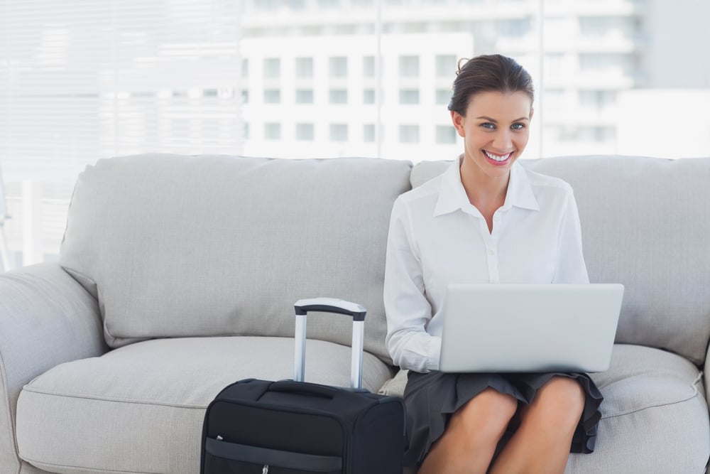 Happy businesswoman sitting on the couch using laptop with her suitcase beside her